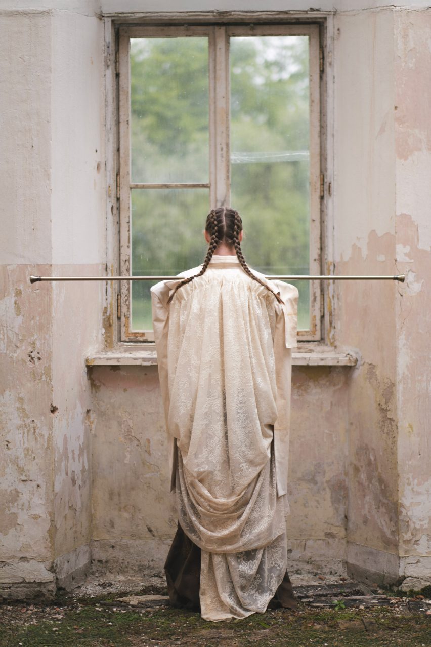 a photograph of a person from behind, standing in front of a window wearing a beige piece of fabric