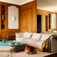 Photograph of the living space, showing a modular white sofa. There is a small dog in the photo.