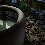 A close-up photograph of a circular, stone bathtub in an outdoor courtyard garden which features plants and a bamboo wall.