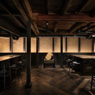 A warmly lit dining area with a dark wooden floor, ceiling and dining tables. A long dining table takes up the left side of the photo. Two shorter dining tables make up the right side of the photo. A small sculpture is in the back, centre of the image.