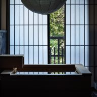 A photo of a large, square, wooden bathtub and behind it, latticed sliding doors made from washi-paper are slightly open to show the outdoor greenery.