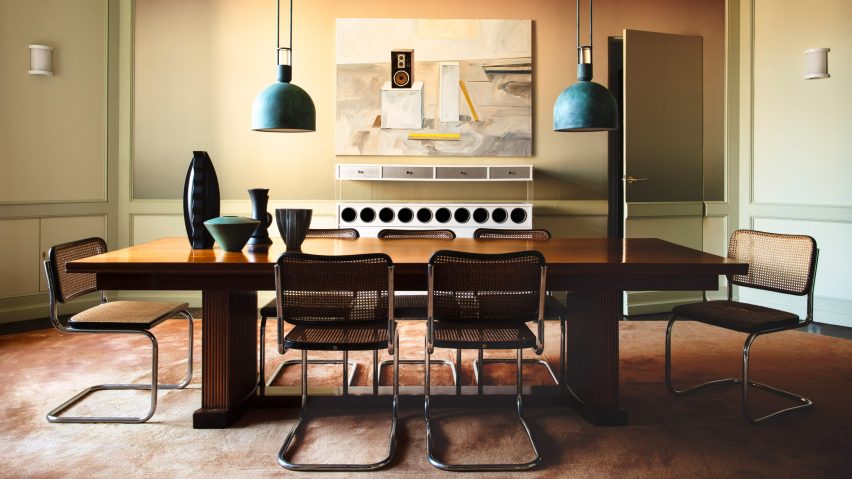 Photograph of the mid-century inspired dining space, showing a large central dining table, with eight chairs and two blue, low-hanging pendant lights. There is artwork on the back wall and a large stereo system.