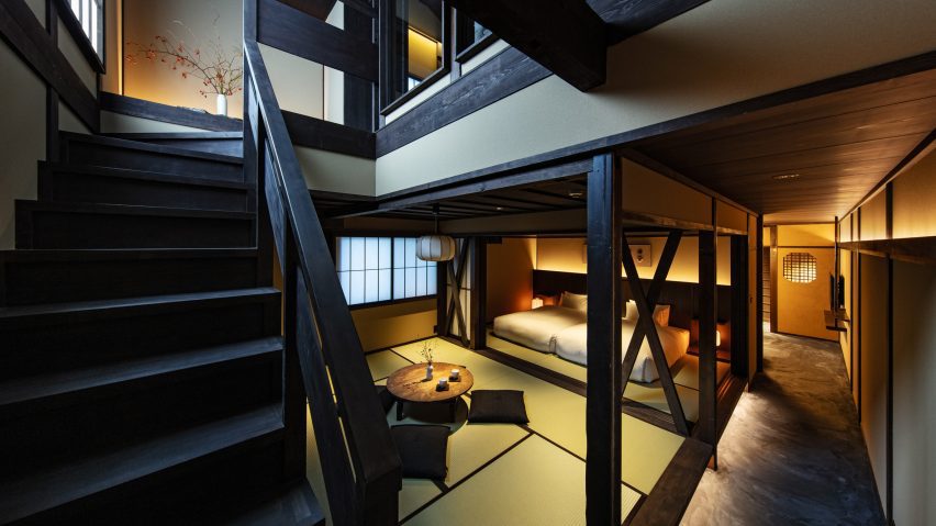 A wooden staircase and banister go up the left side of the photo, with a vase and flowers at the top of the stairs. A warmly lit, open plan area contains a small coffee table with futon-cushions for sitting and two beds towards the back of the space.