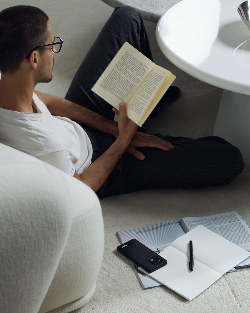 Photo of a casually dressed man facing away from the camera as he relaxedly sprawls and reads a book, the Punkt MC03 phone sitting facedown on top of an open notebook at his side.
