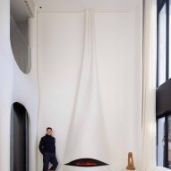 A tall, double-height fireplace that is integrated into a wall that has been carved to resemble a piece of draped cloth. The main architect, Nelson Chow is standing on the left of the fireplace with a dog laid on the floor beside him.