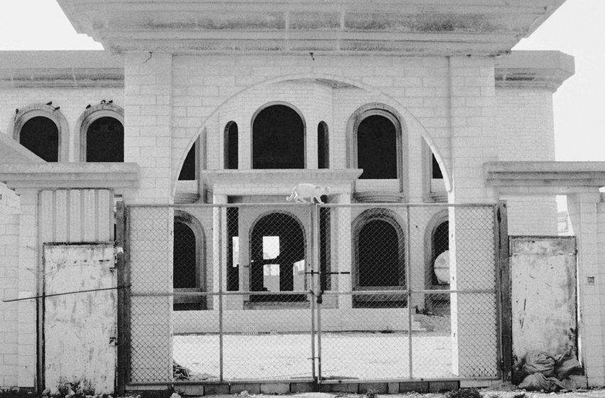 Black and white photograph of a cat on a metal gate, with a large arched building in the background.