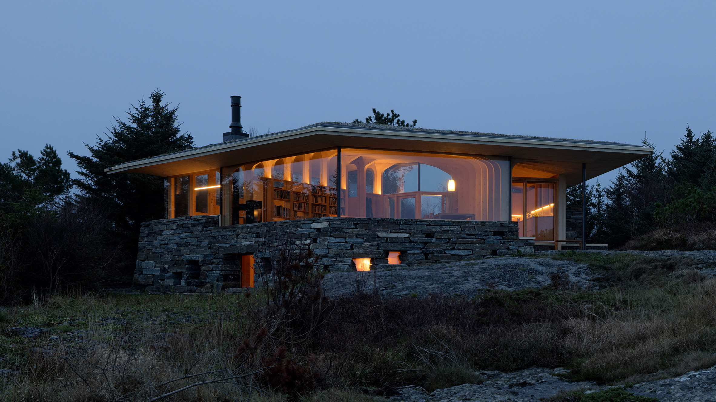 Evening view of the illuminated timber cabin resting on a stone base within the coastal landscape