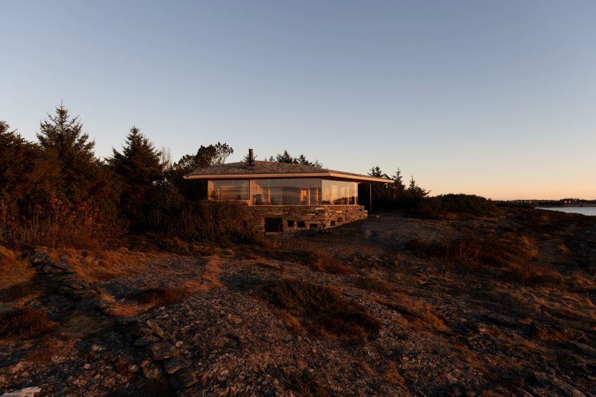 Exterior view of the low timber cabin on a rocky coastal site at sunset