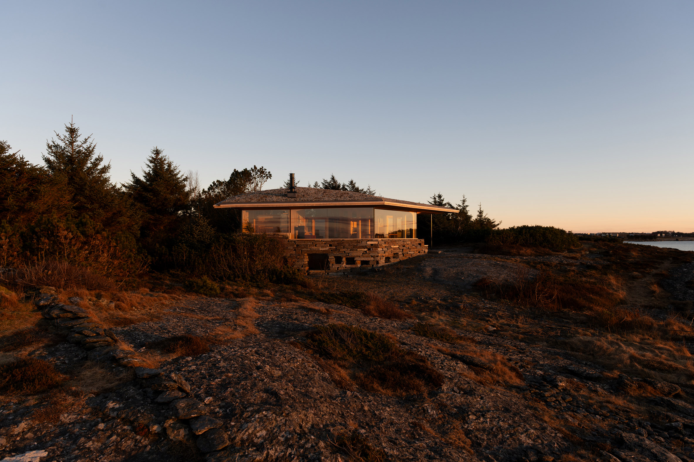 Exterior view of the low timber cabin on a rocky coastal site at sunset