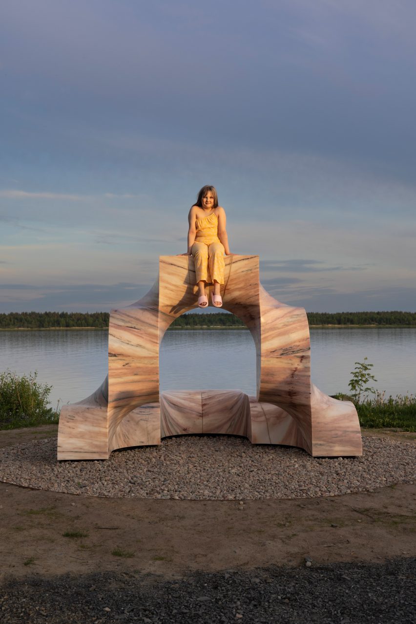 Photo of a girl smiling from atop of the pink arched marble Super Kello sculpture in front of still harbour waters