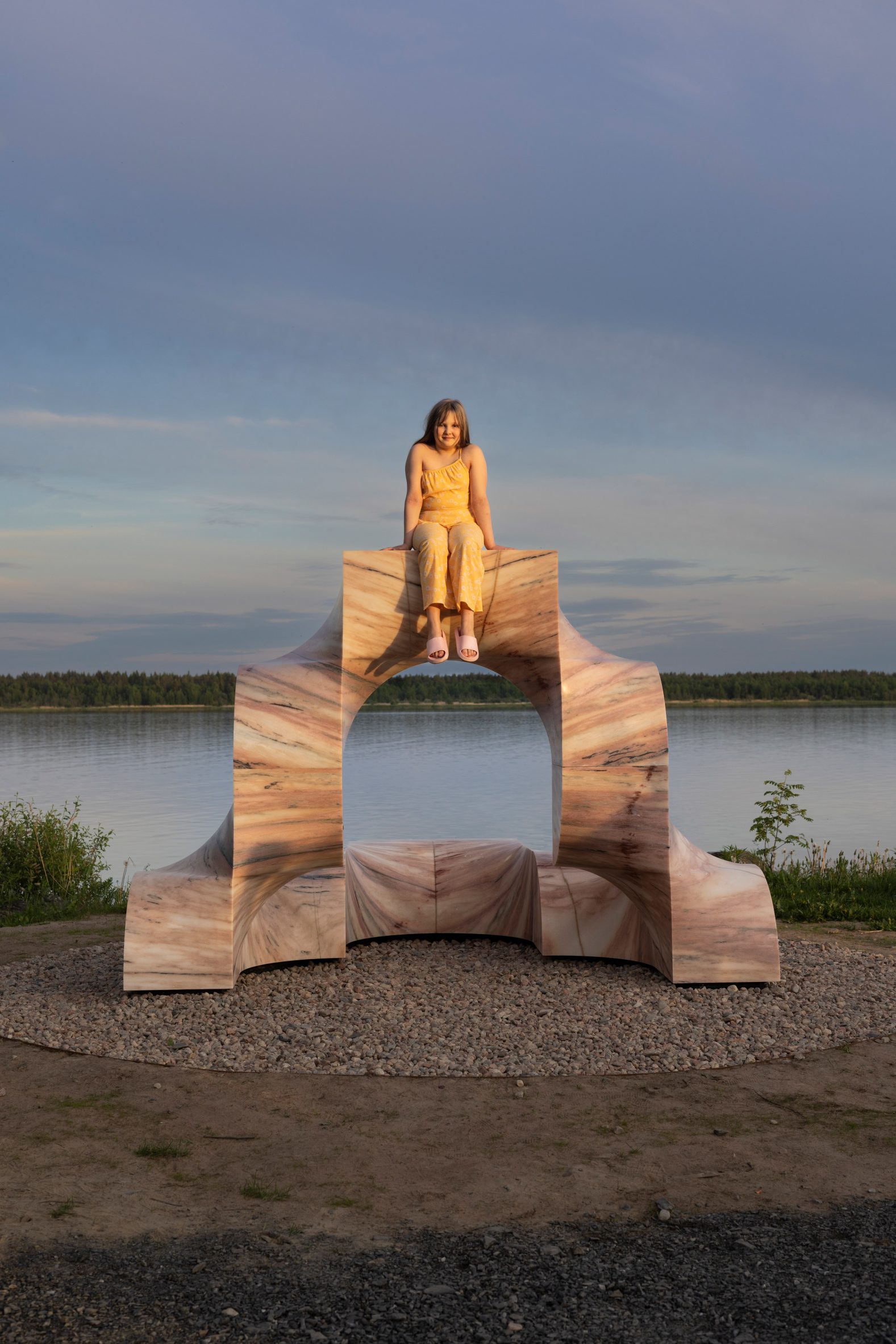 Foto de una niña sonriendo encima de una escultura arqueada de mármol rosa Super Kailo frente a las tranquilas aguas del puerto