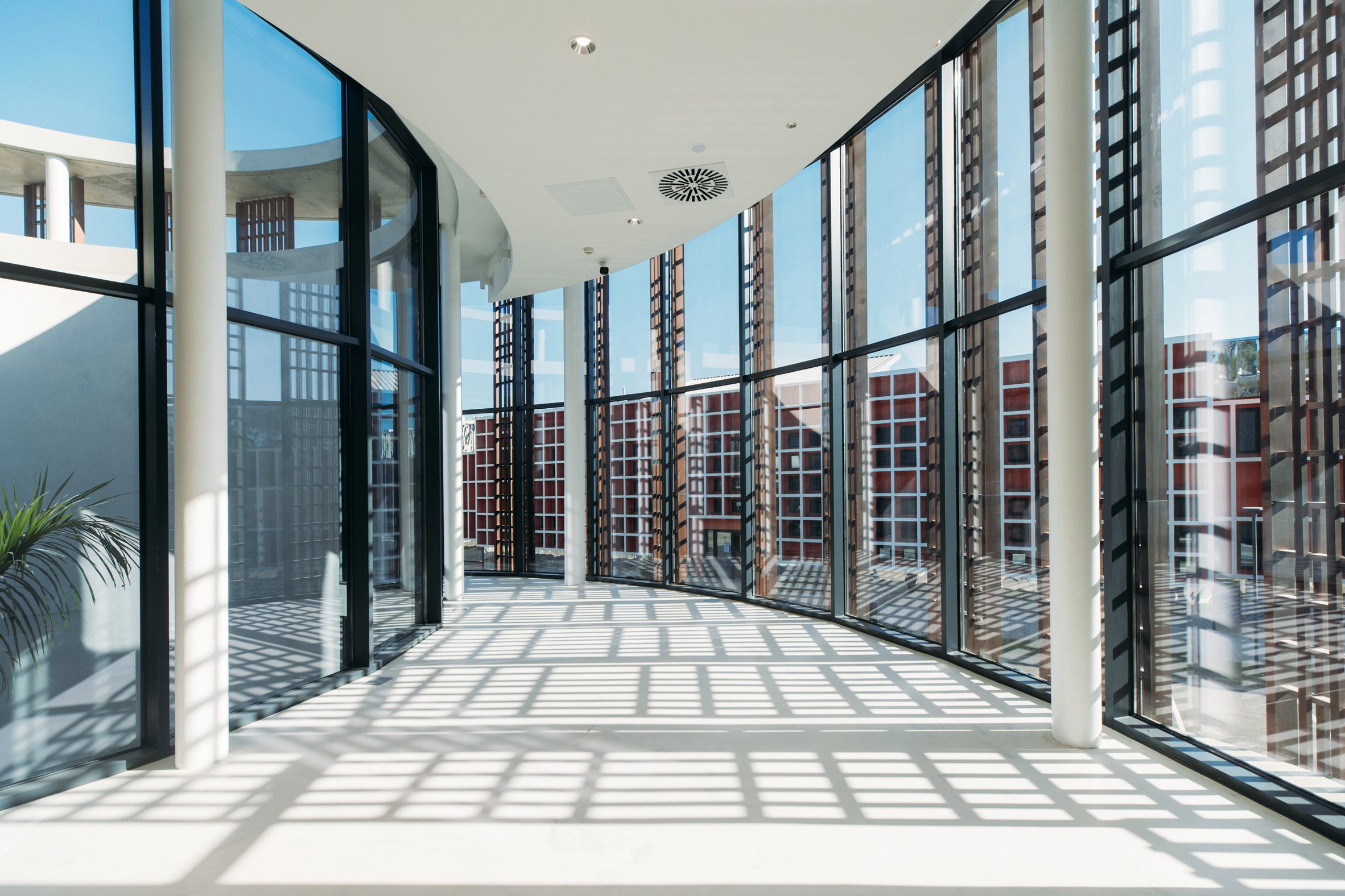 Curved interior corridor with full-height glazing and patterned shadows cast across the floor