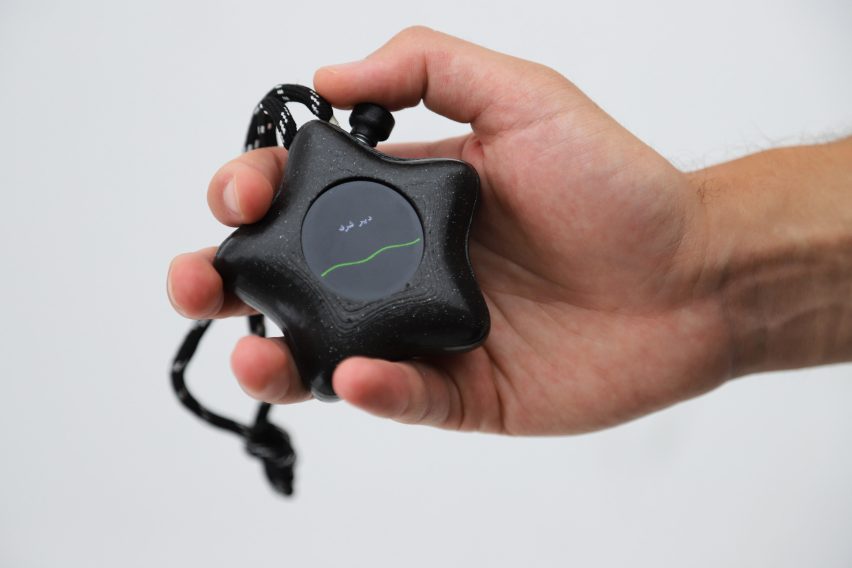 Photo of a hand holding the Mesh-89 device with a slightly wavy green line across a circular black screen with Arabic text above it 