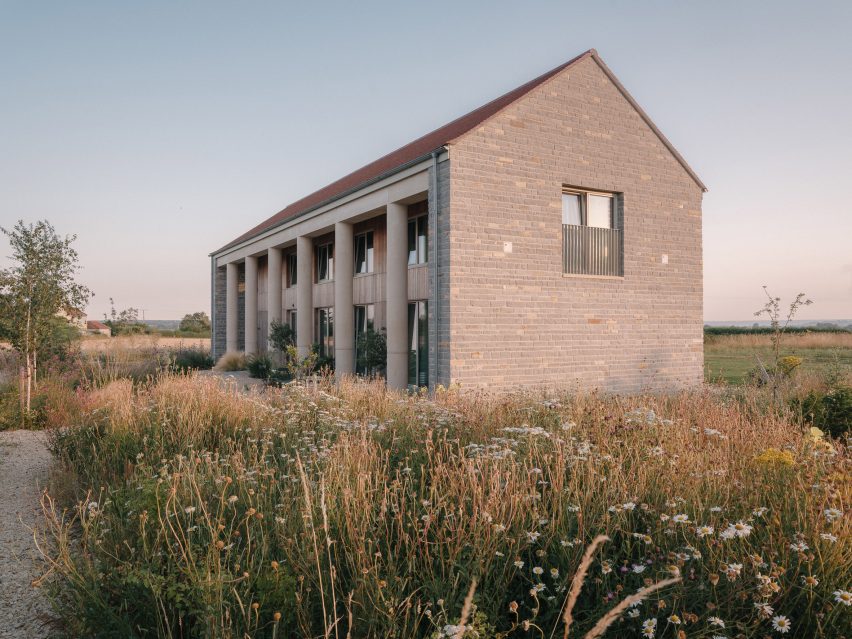 View across meadow grasses towards the rear of the house, showing its pitched roof, stone walls and lower wing extending into the landscape