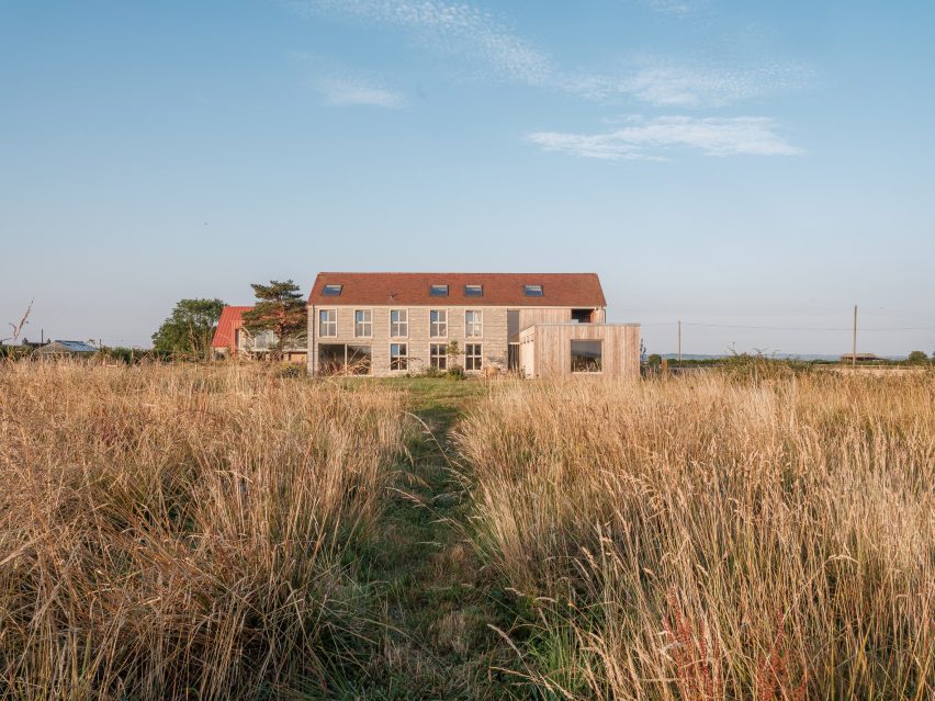 View across meadow grasses towards the rear of the house, showing its pitched roof, stone walls and lower wing extending into the landscape