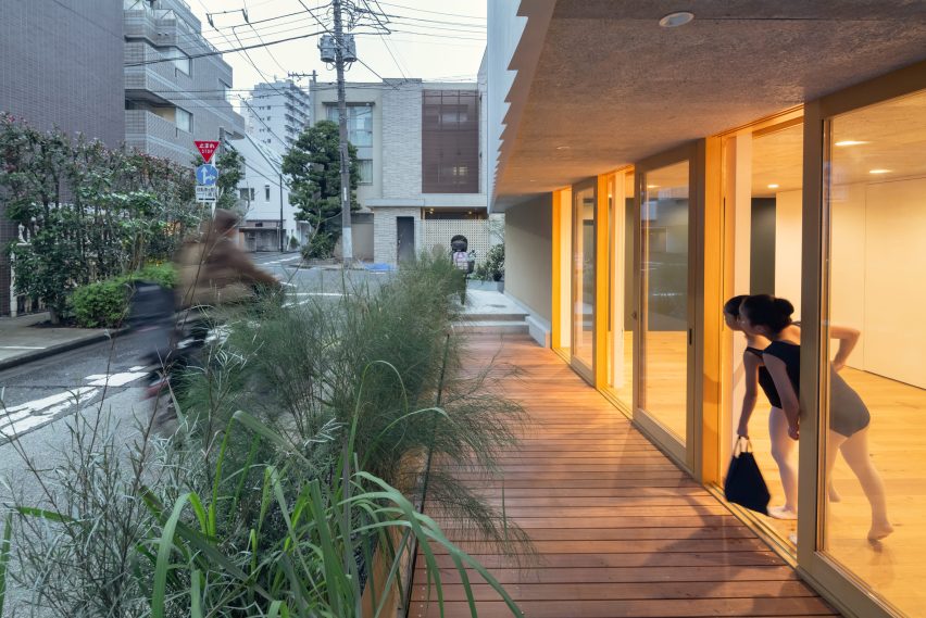 Ballet students stand beside a glazed facade facing the street, with timber decking and planting between the building and pavement