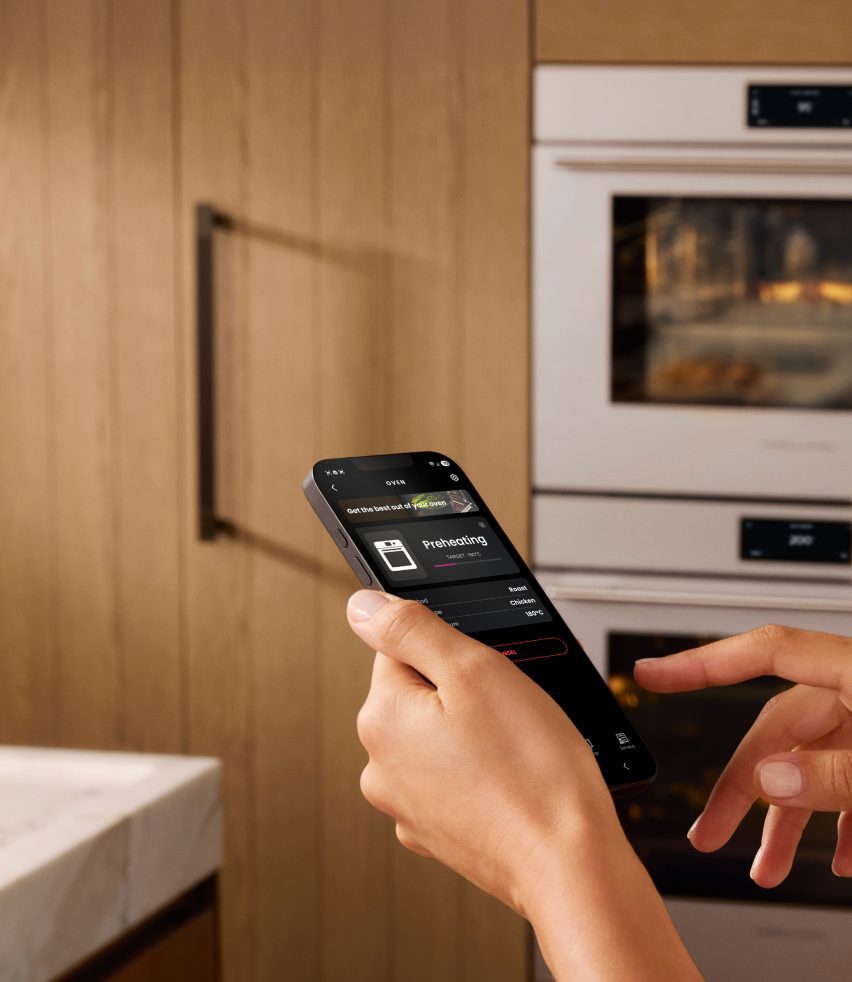 Close-up image of a person's hands while they hold their phone and look at an app that tells them their oven is preheating while they stand in their kitchen