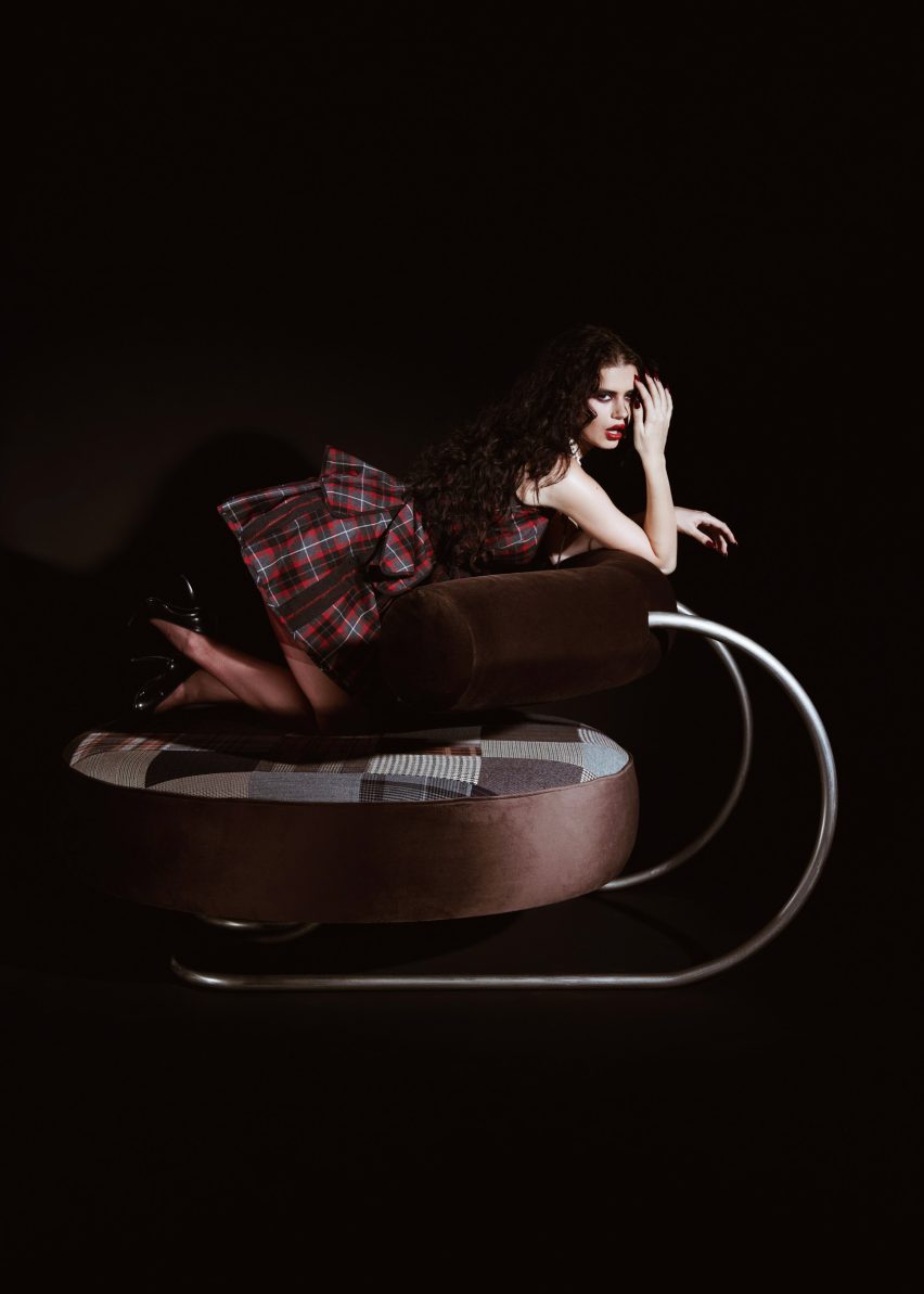 Photograph of a person kneeling on a large circular seat, leaning on the curved cylindrical backrest of the seat against a dark background. The image shows the side profile of the chair and the exaggerated metal frame.