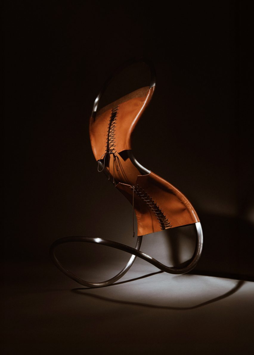 A chair made from a continuous, tubular, stainless steel frame covered with tanned leather and secured with brown cord that has been laced like a corset. The chair is leant forward and to the left and looks like it is precariously balanced.