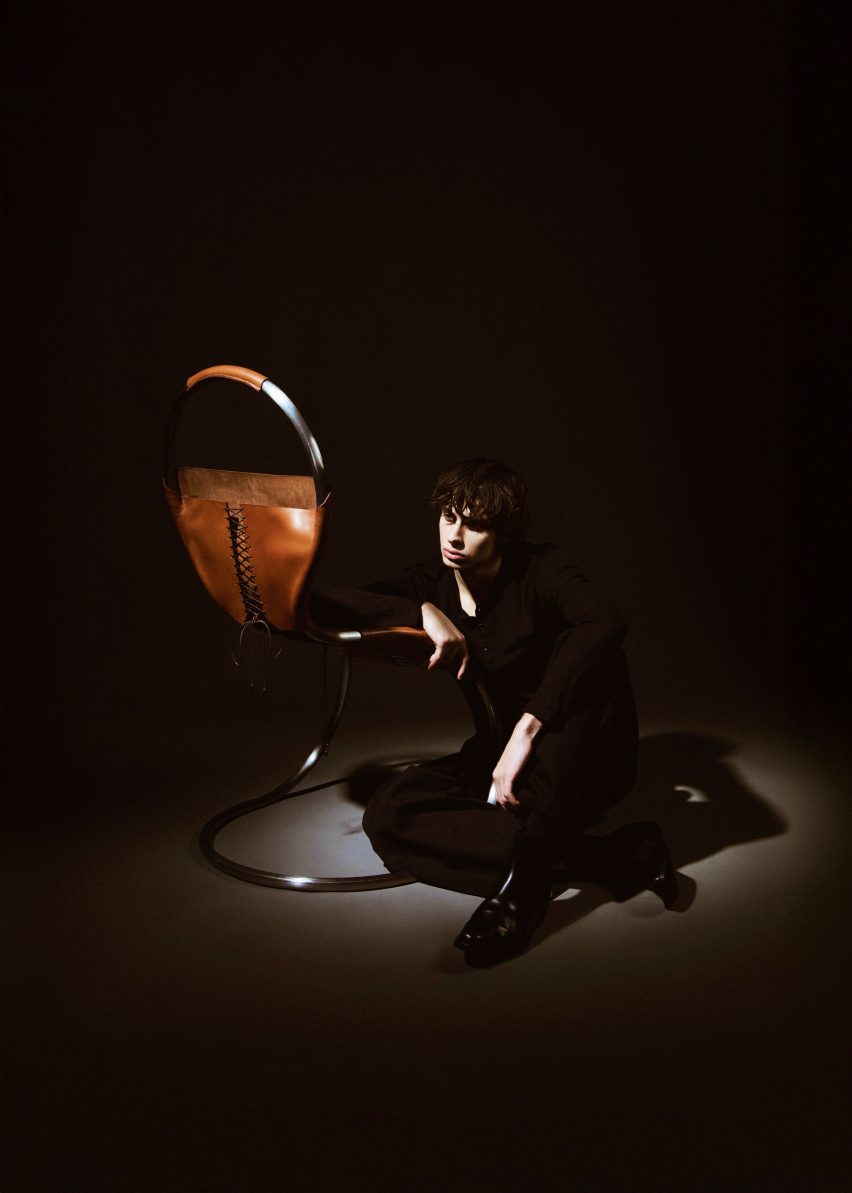Image of a person sitting on the floor and leaning on a chair made from tanned leather that has been wrapped around a stainless steel frame. The leather has laced detailing resembling a corset. 