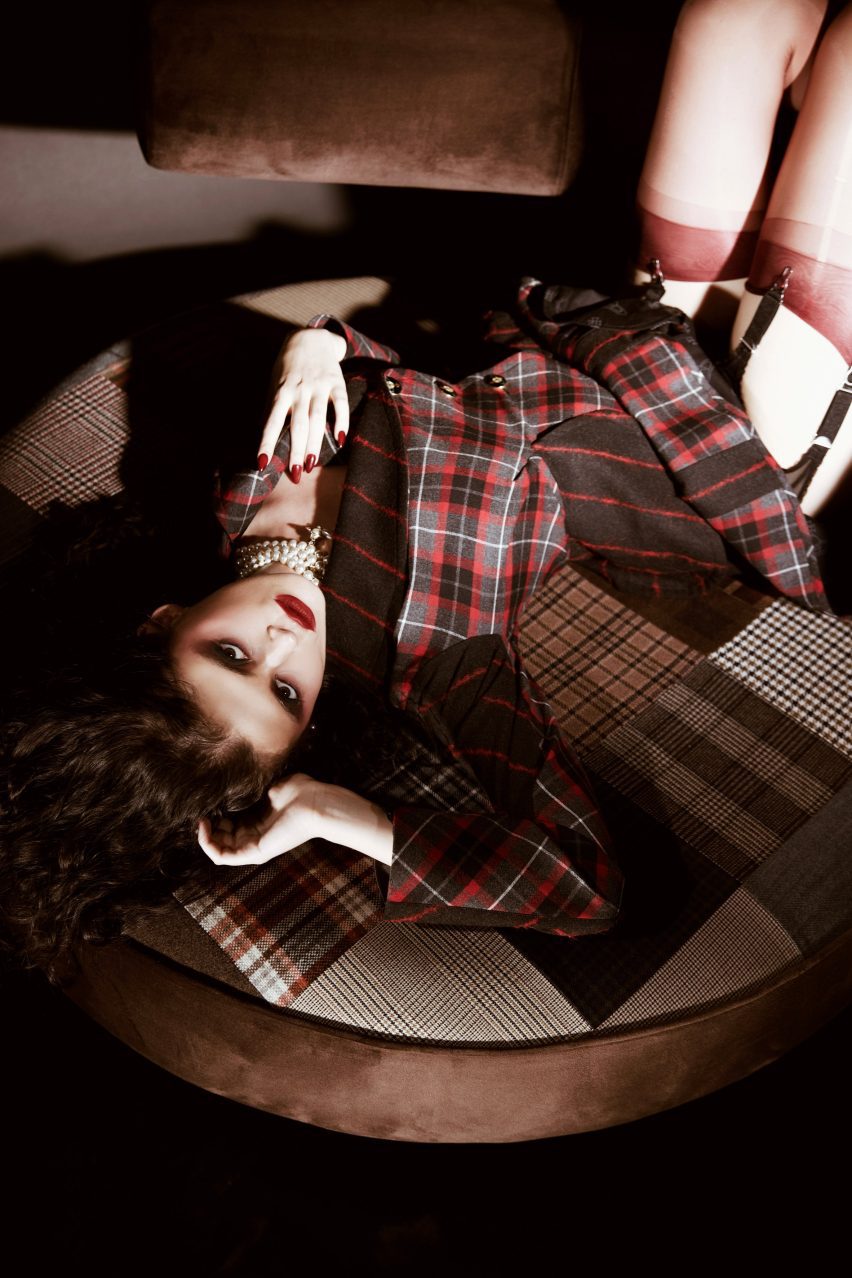 Photograph of a woman dressed in tartan-patterned clothing lays across a large circular seat. The seat features patchwork upholstery.
