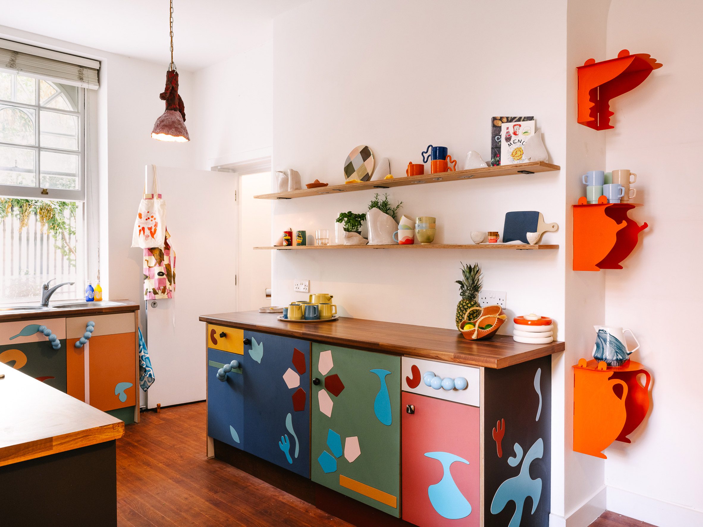 A photo of the kitchen at the UAL Atterbury Flat with white walls and colourful cabinetry inlaid with bright and bold contemporary marquetry, two open shelves over the kitchen bench lined with ceramics and quirkly kitchen tools, bright orange metal shelves in a nook with cut-out vessel shapes when viewed in profile, and other colourful details