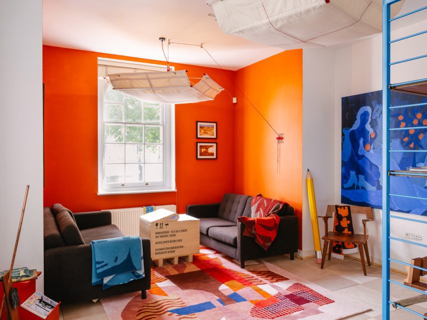 Photo of the living room of the UAL Atterbury Flat showing a space cocooned in orange walls, with a coffee table made from a stack of paper disguised as a packing crate, a colourful rug, and a floaty pendant light that looks like a vast canvas canopy supported by the faint outlines of an internal structure