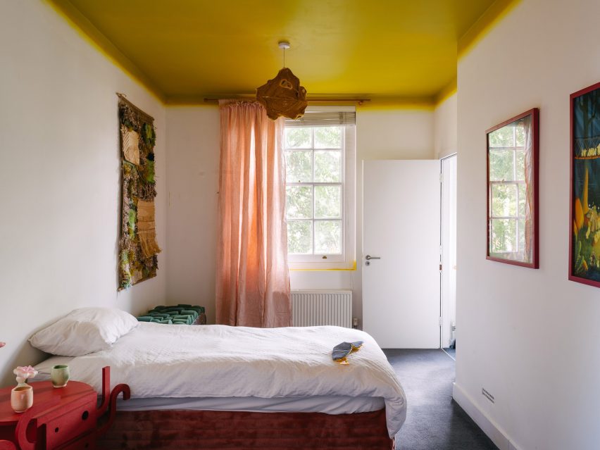 Photo of the "whimsical" bedroom at the UAL Atterbury Flat, featuring a chartreuse-coloured ceiling with a neon gradient effect as it merges into white walls, soft pink curtains, a shaggy textile work on one wall and a bedside table in the foreground that looks like an abstracted alien character