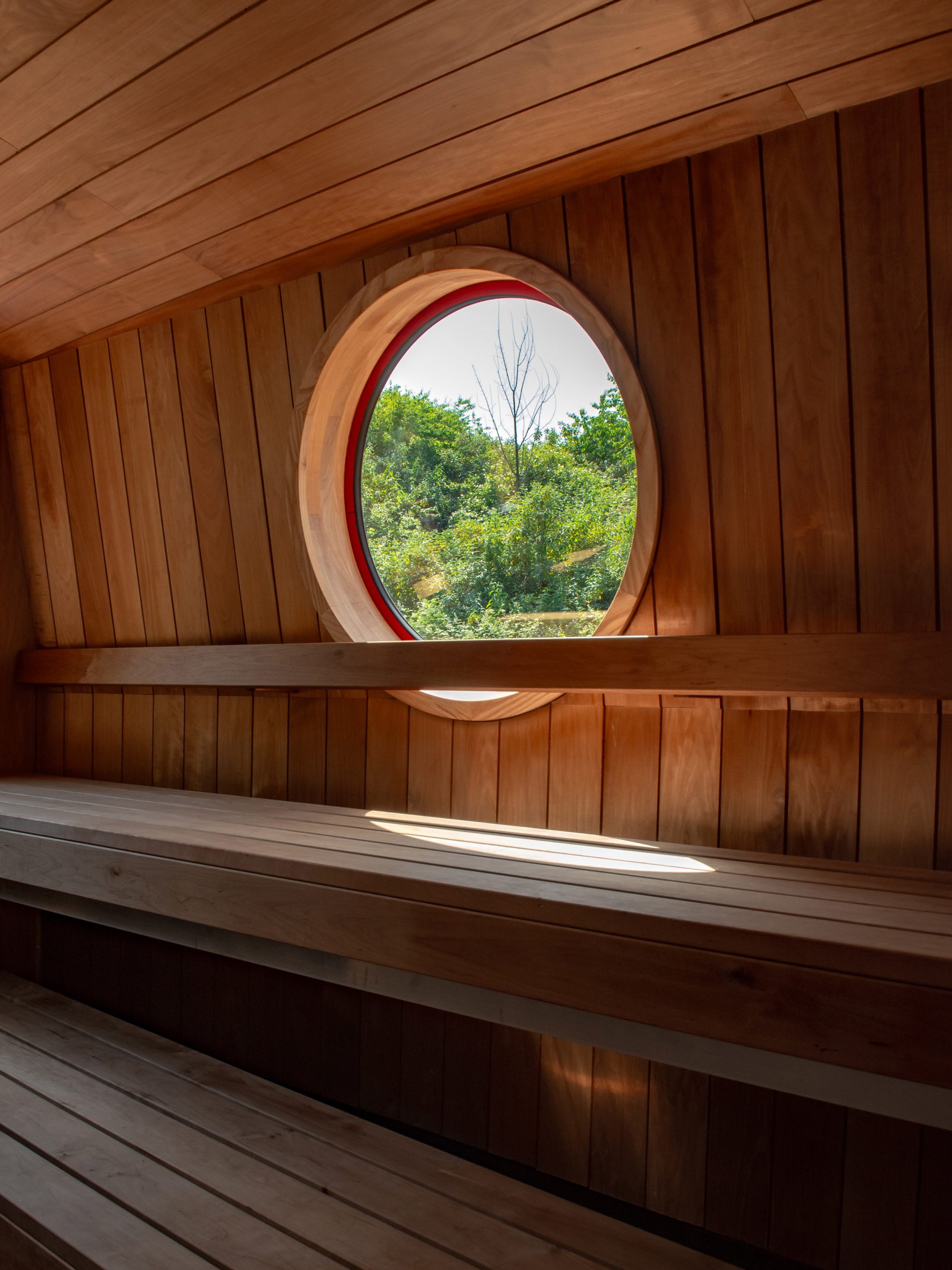 Timber sauna bench beneath a circular porthole overlooking trees
