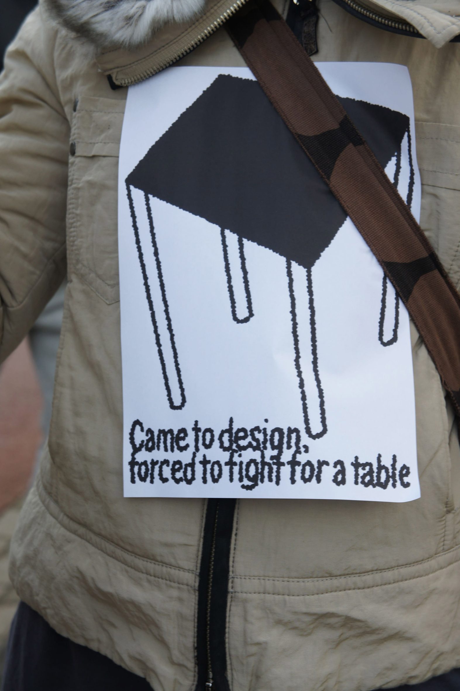 Close-up photo of a protest sign stuck to a person's jacket reading Came to design, forced to fight for a table